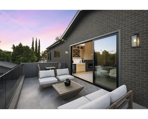 Moderne Terrasse mit Fliesenboden, Lounge Möbeln und Glasgeländer vor einem Haus mit dunkler Ziegelsteinfassade bei Abenddämmerung.