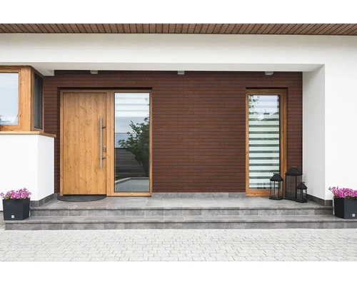 Eingangsbereich eines Hauses mit einer Holztür, Seitenteil aus Glas, Ziegelsteinwand und gefliester Treppe mit Pflanzgefäßen und Laternen.