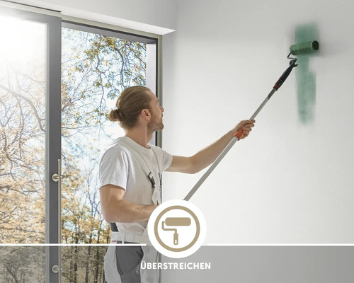 Ein Maler streicht eine Wand mit einer Farbrolle und einem Teleskopstab, Symbol für das Überstreichen von Wandflächen.