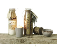 Deux bouteilles en verre avec préparations pour gâteaux, une bougie dans un pot, une bobine de fil et deux petits bols sur une planche en bois.
