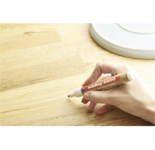 Une main utilise un marqueur pour meubles Edding 8900 pour retoucher des rayures sur une surface en bois.