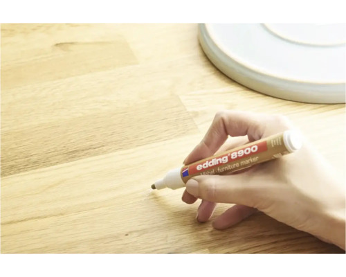 Une main utilise un marqueur pour meubles Edding 8900 pour retoucher des rayures sur une surface en bois.