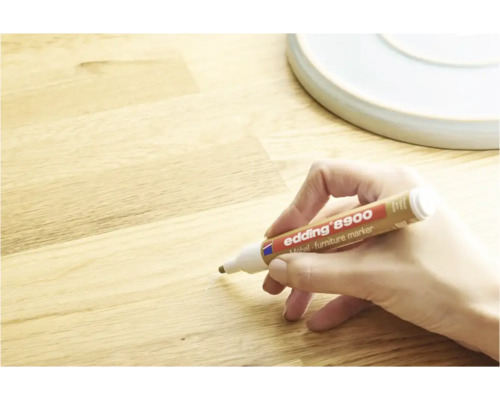 Une main utilise un marqueur pour meubles Edding 8900 pour réparer une rayure sur une surface en bois.