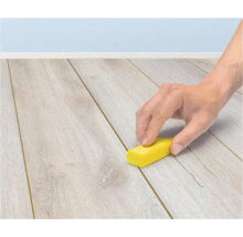 Eine Hand verwendet einen kleinen gelben Block, um eine Fuge zwischen den Dielen eines hellen Holzlaminatbodens zu reinigen.