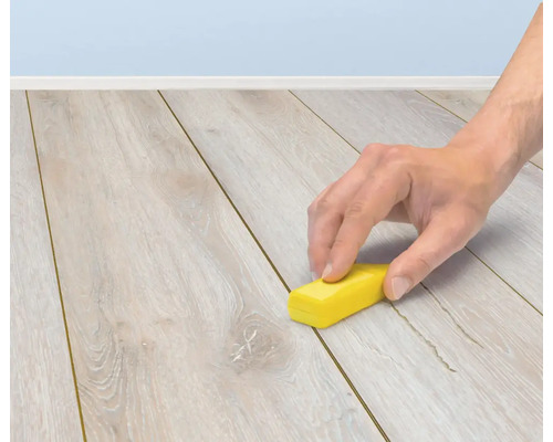 Eine Hand verwendet einen Reparaturwachsstift zur Ausbesserung von Kratzern auf einem Holzlaminatboden.