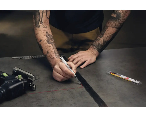 Eine Person zeichnet mit einem Edding Tieflochmarker eine Linie an einem Metalllineal auf einer dunklen Arbeitsplatte.