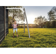 Enfant se balançant sur une balançoire en bois dans le jardin, fixée à une maison en bois noire, au coucher du soleil.