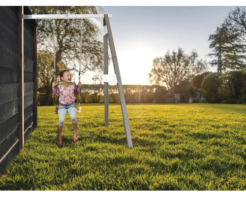 Enfant se balançant sur une balançoire en bois dans le jardin, fixée à une maison en bois noire, au coucher du soleil.