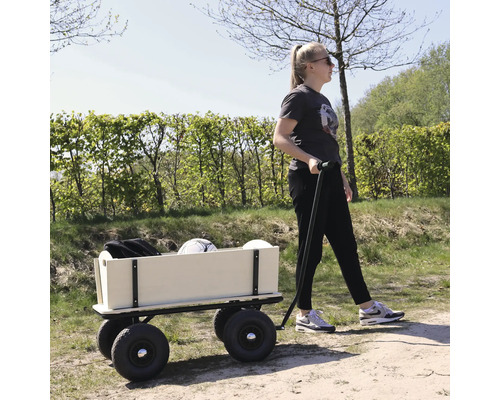 Une femme tire une charrette en bois avec des roues en caoutchouc sur un chemin extérieur par temps ensoleillé.