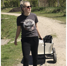 Une jeune femme tire une charrette en bois avec des roues en caoutchouc sur un chemin de terre en extérieur.