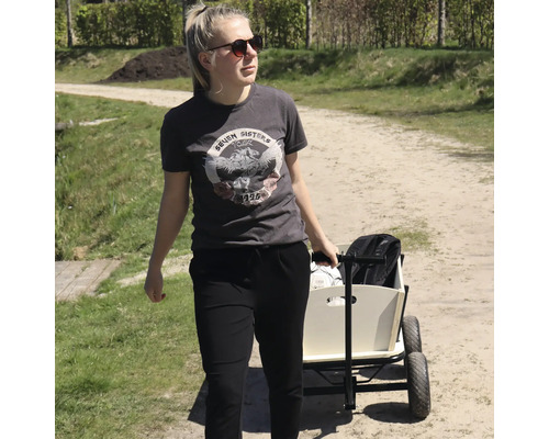 Une jeune femme tire une charrette en bois avec des roues en caoutchouc sur un chemin de terre en extérieur.