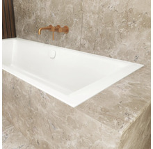 Baignoire encastrable blanche dans une salle de bain avec revêtement en marbre beige et un mitigeur mural de couleur cuivre.