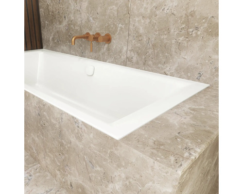 Baignoire encastrable blanche dans une salle de bain avec revêtement en marbre beige et un mitigeur mural de couleur cuivre.