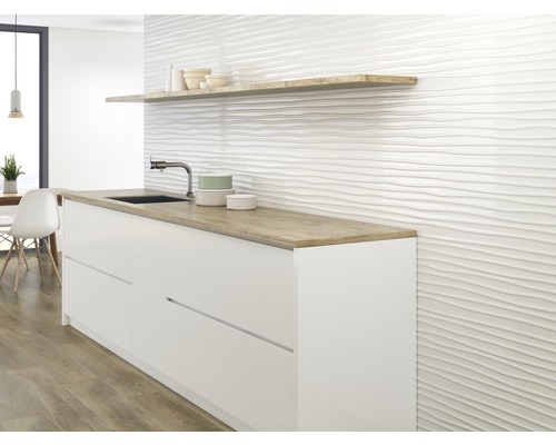 Küchenszene mit weißen Unterschränken, einer Holzablage und einer Wand mit gewellter Fliesenstruktur.