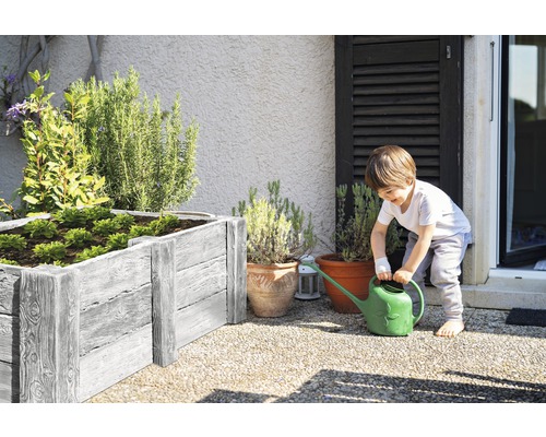 Un garçon arrose des plantes dans un potager surélevé et des pots de fleurs dans le jardin.