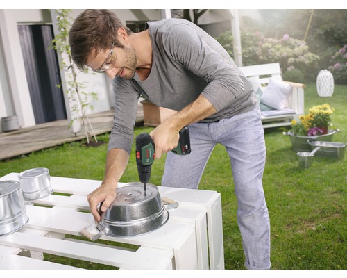 Un homme perce des trous dans un seau en métal dans le jardin avec une perceuse-visseuse sans fil.