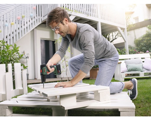 Un homme travaille avec une visseuse sans fil sur un projet de bricolage à partir de palettes dans le jardin.