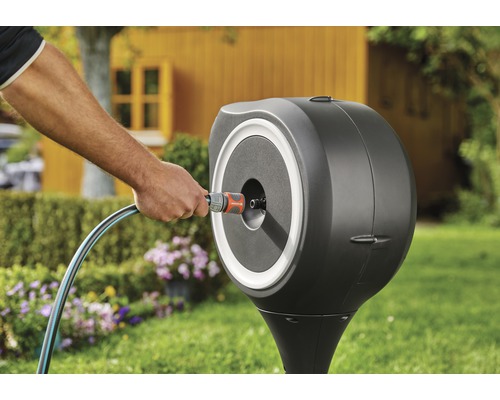 Gartenszene mit Schlauchbox an Rasenfläche mit Blumen und Gartenhaus im Hintergrund