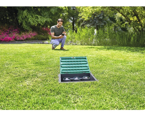 Gartenlandschaft mit einem Mann, der ein Smartphone bedient, im Vordergrund ein im Rasen eingelassener Stromverteilerkasten mit geöffnetem Deckel.