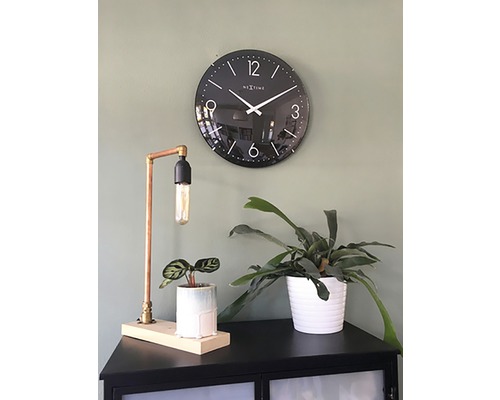 Schwarze Wanduhr neben Tischleuchte und Zimmerpflanzen auf einem Schrank