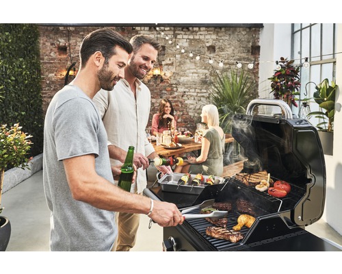 Zwei Männer grillen Fleisch und Gemüse auf einem Gasgrill im Garten, während sich andere an einem Holztisch unterhalten.
