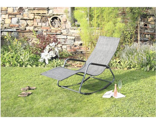 Gartenszene mit einer grauen Schaukelliege, einer Flasche, einem Weinglas, einem Buch und Sandalen auf dem Rasen vor einer Steinmauer mit Insektenhotel.