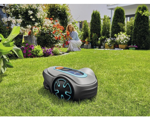 Rasenmähroboter auf einer Rasenfläche mit einer Frau im Hintergrund, die sich um Blumen kümmert.
