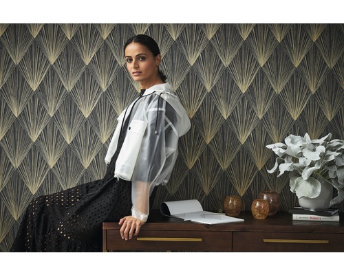 Szene mit gemusterten Tapeten und einer Frau, die auf einem Schreibtisch sitzt, auf dem eine Pflanze, Vasen und Bücher stehen.