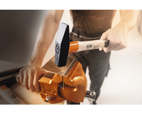 Ein Mann bearbeitet ein Metallstück mit einem Hammer und einem Schraubstock.