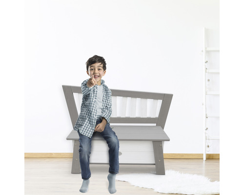 Un garçon est assis sur un banc coffre à jouets gris et pointe du doigt.