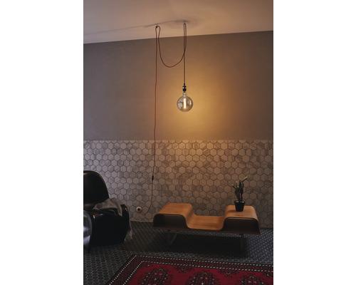 Innenaufnahme eines Wohnzimmers mit einer einzelnen Glühbirne als Pendelleuchte, einem Sessel, einem Holztisch und einem roten Teppich.