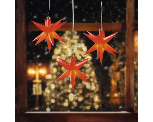 3er-Set Sternenhänger Ø 12 cm 3 LEDs Lichtfarbe warmweiß rot Drei rote Sterne als Fensterdekoration vor verschneiter Landschaft und Weihnachtsbaum