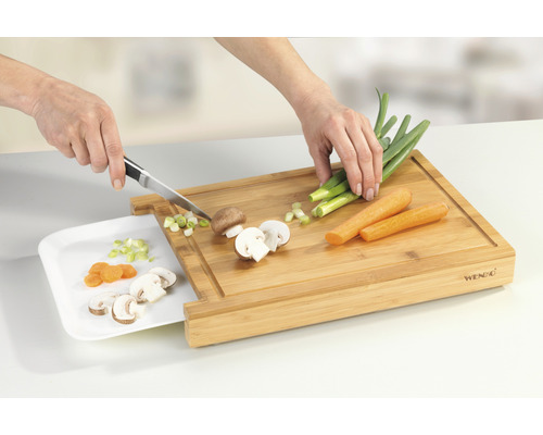 Person schneidet Gemüse auf einem Bambus Schneidebrett mit Schublade für Abfälle.