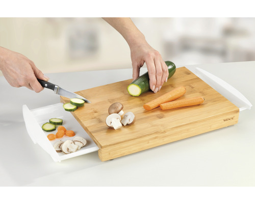 Bambus Schneidebrett mit Auffangschale, darauf Zucchini, Karotten und Pilze, die von einer Person mit einem Messer geschnitten werden.