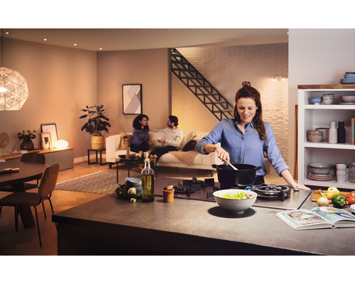 Helle, einladende Küche mit Frau, die am Herd kocht, und einer entspannten Familienszene im Hintergrund.
