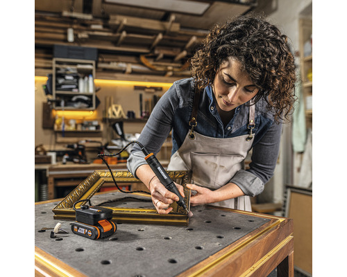 Une femme travaille avec un outil multifonction sans fil Worx sur un cadre photo doré dans un atelier.