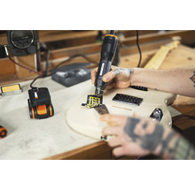 Eine Person entfernt mit einem Heißluftgebläse einen Aufkleber von einer Gitarre. Eine Kabeltrommel steht auf dem Tisch.