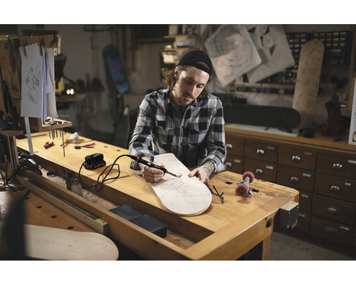 Mann arbeitet mit einem Lötkolben an einem Skateboard in einer Werkstatt.