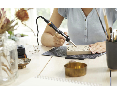Frau arbeitet mit einem Brennkolben an einem Holzstück auf einem Schreibtisch.