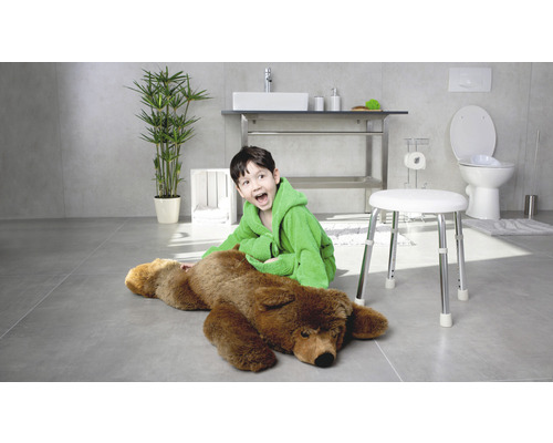 Salle de bains lumineuse avec un garçon en peignoir assis à côté d''un ours sur un tabouret, avec lavabo, toilettes et plante