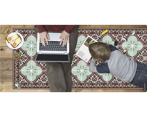 Ein Zimmer mit einem Teppich im Fliesenmuster, auf dem eine Person an einem Laptop arbeitet und ein Kind liegt.