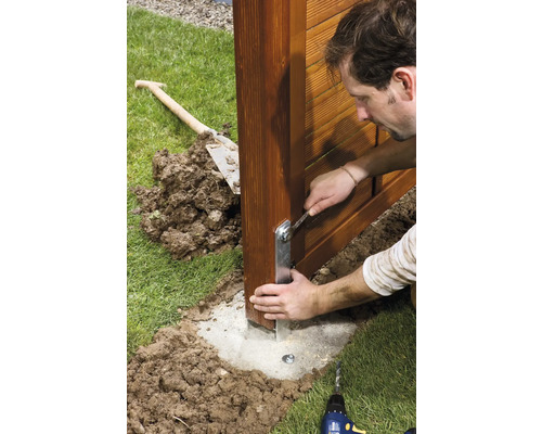 Mann montiert Zaunpfosten mit Schraubenschlüssel auf Betonfundament im Garten.