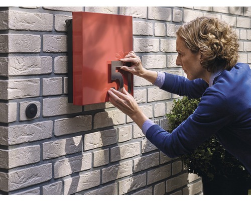 Frau befestigt eine Hausnummer an einem roten Briefkasten an einer Ziegelmauer