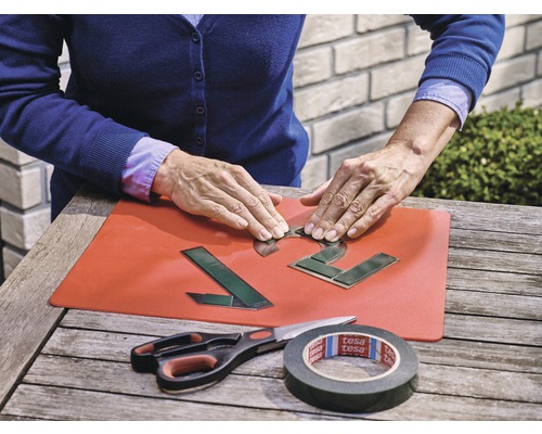 Eine Person befestigt Klebeband an Hausnummern auf einer roten Unterlage, mit einer Schere und einer Tesa Klebebandrolle auf einem Holztisch.