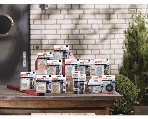 Présentation de diverses solutions de collage Tesa sur une table en bois devant un mur de briques et un buisson vert.