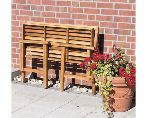 Zusammengeklappte Gartenbank aus Holz neben einem Blumentopf mit roten Blüten vor einer Backsteinmauer.