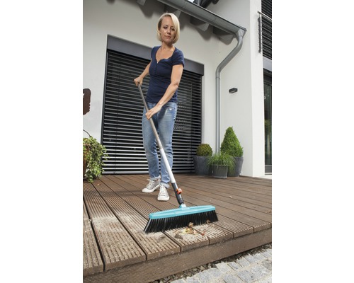 Une femme nettoie une terrasse en bois avec un balai de voirie.