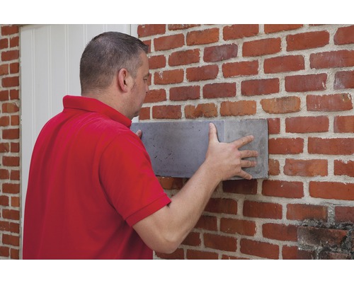 Un homme fixe un bloc de béton sur un mur en briques.
