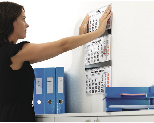 Frau befestigt einen Kalender an der Wand im Büro neben Ordnern und Ablagekörben.
