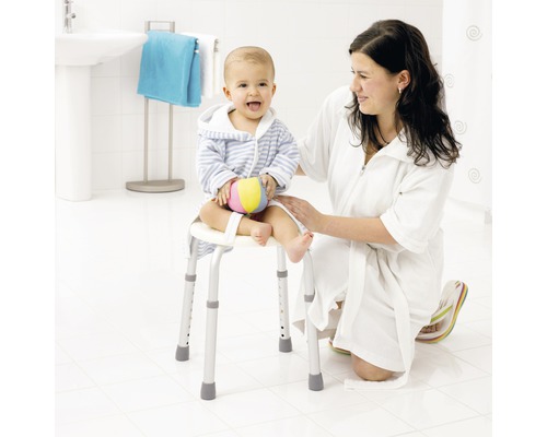 Eine Frau kniet neben einem Kind, das auf einem Duschhocker mit Armlehnen sitzt, in einem hellen Badezimmer.